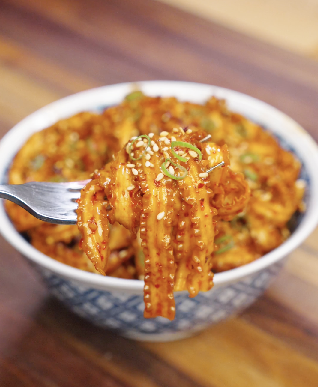 Fork lifting a bite of garlic chili noodles coated in spicy sauce with sesame seeds and scallions.