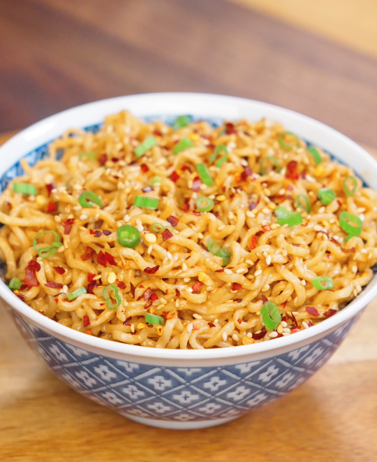 Bowl of garlic sesame noodles tossed in creamy sesame sauce and topped with chili flakes, sesame seeds, and sliced scallions.