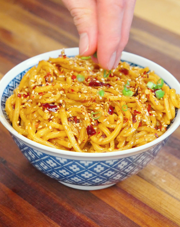 Hand sprinkling sliced scallions over a bowl of chili oil garlic butter noodles topped with sesame seeds and chili flakes.