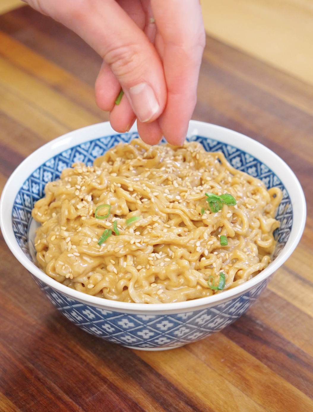 Hand sprinkling scallions and sesame seeds over a bowl of creamy peanut noodles.
