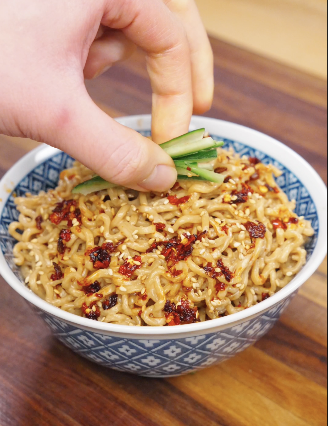 Hand placing sliced cucumber garnish on top of creamy sesame noodles with chili oil and sesame seeds.