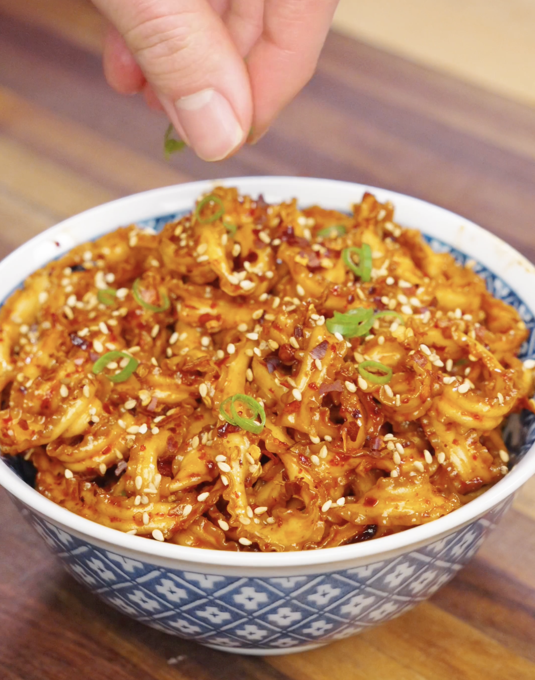 Hand sprinkling sliced scallions over a bowl of garlic chili noodles topped with sesame seeds and chili flakes.