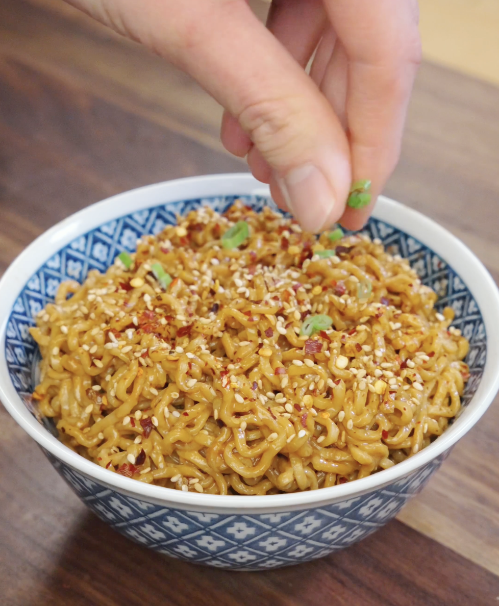 Fresh scallions being sprinkled over garlic peanut butter noodles finished with sesame seeds and chili flakes.