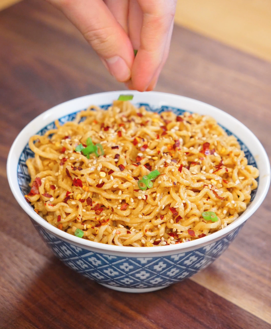 Hand sprinkling sliced scallions over a bowl of garlic sesame noodles topped with chili flakes and sesame seeds.
