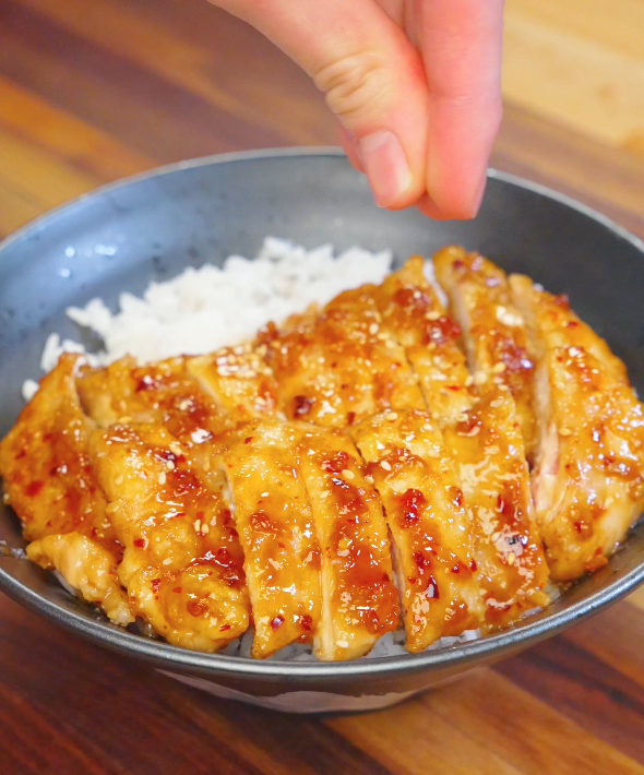 Hand sprinkling sesame seeds over sliced Honey Chili Chicken served over steamed rice in a bowl.