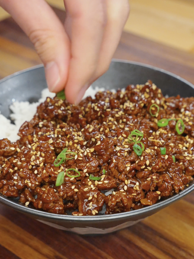 Hand sprinkling sesame seeds and sliced scallions over Korean ground beef recipe in a bowl.