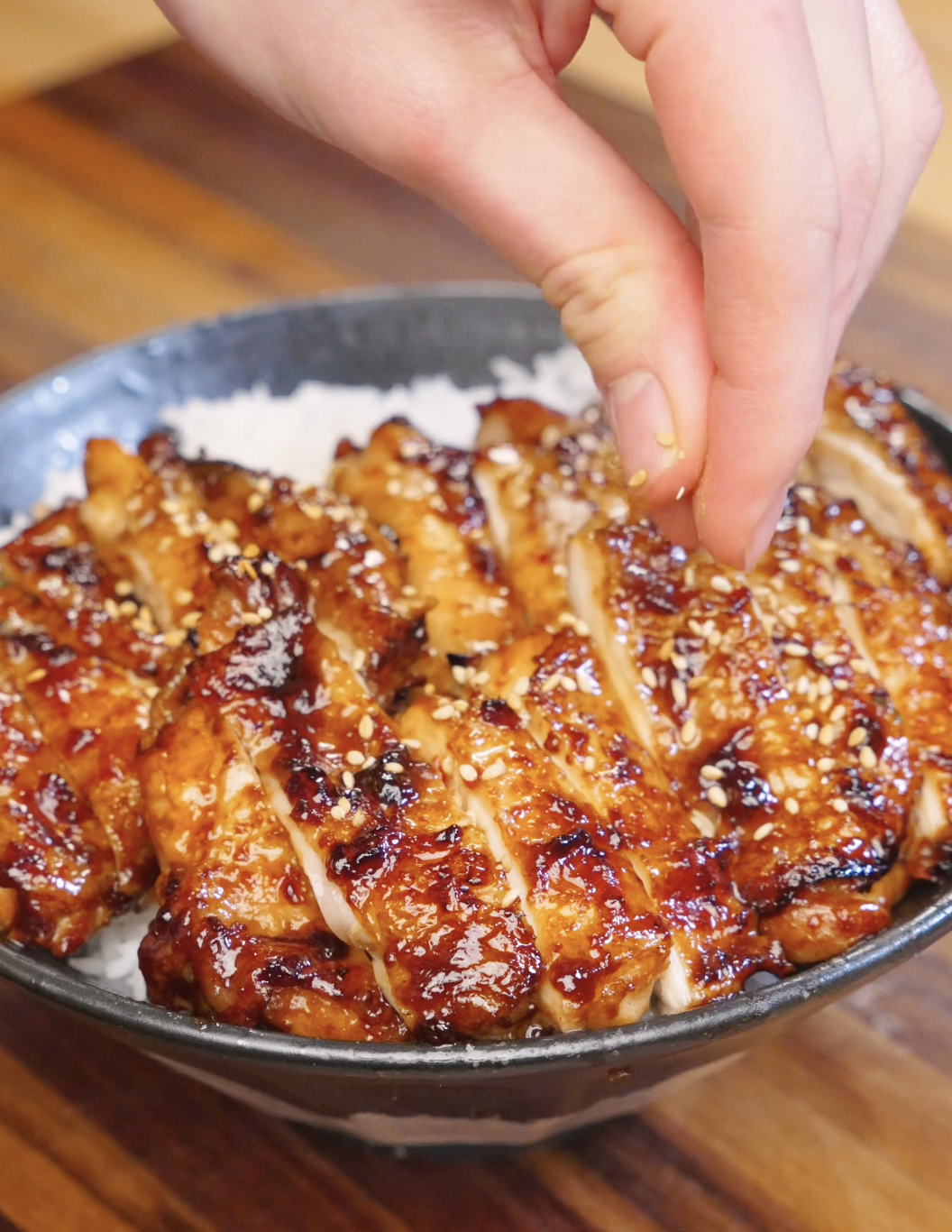 Hand sprinkling sesame seeds over sliced Sweet Chili Chicken served on a bowl of rice.