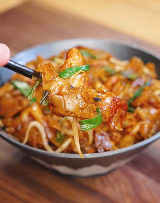 Chopsticks lifting a bite of chicken chow fun with tender chicken, wide rice noodles, bean sprouts, and scallions coated in glossy chow fun sauce.