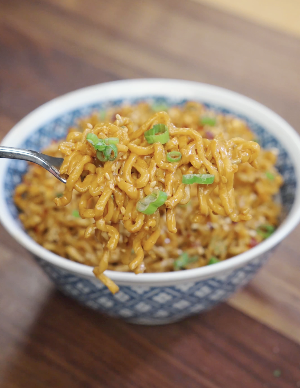 Spoon lifting a bite of creamy garlic peanut butter noodles garnished with scallions and sesame seeds.