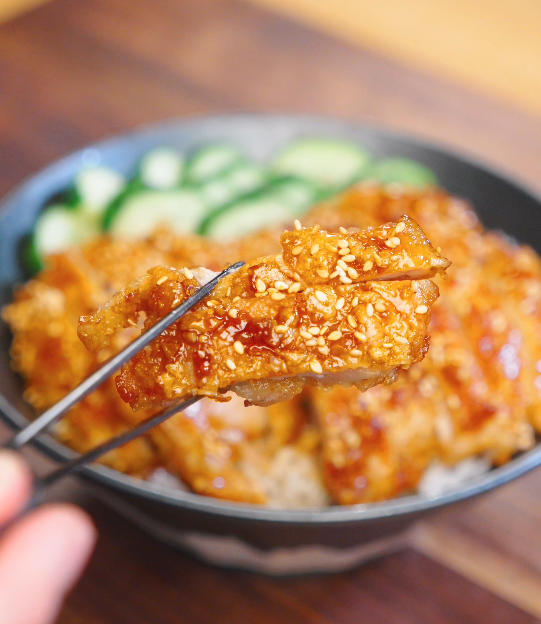 Chopsticks holding a glossy slice of crispy garlic chicken coated in sticky soy garlic glaze and sesame seeds.