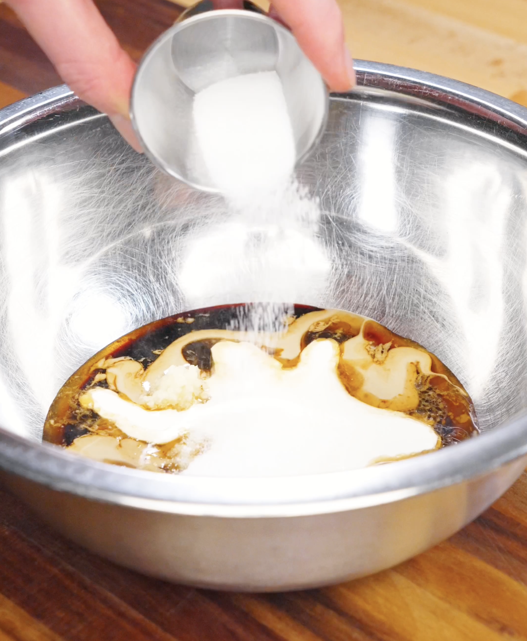 Sugar being poured into a bowl with sesame paste, soy sauce, garlic, and cream to make creamy sesame noodle sauce.