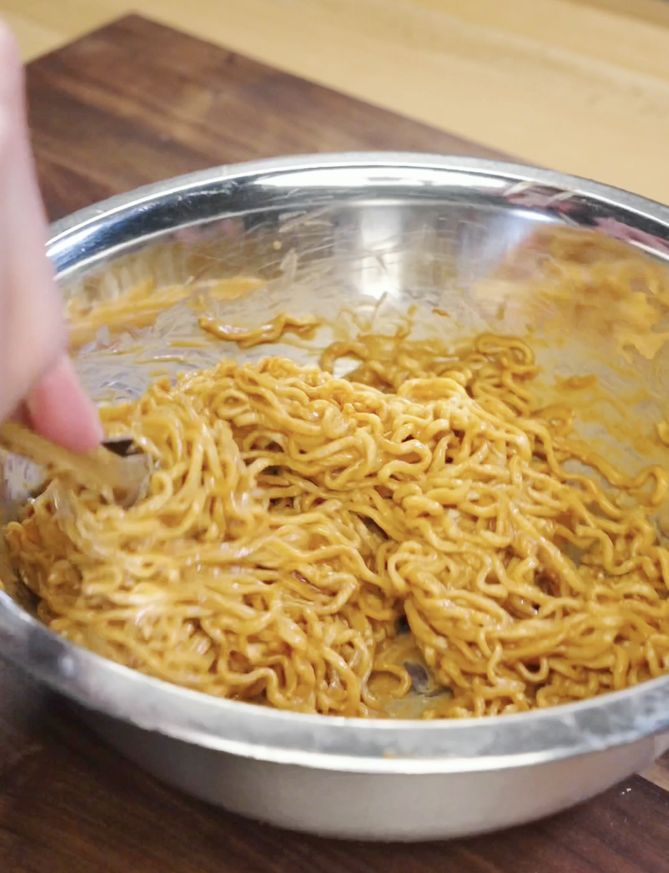 Cooked noodles being tossed in a bowl with creamy peanut butter garlic sauce until every strand is evenly coated.