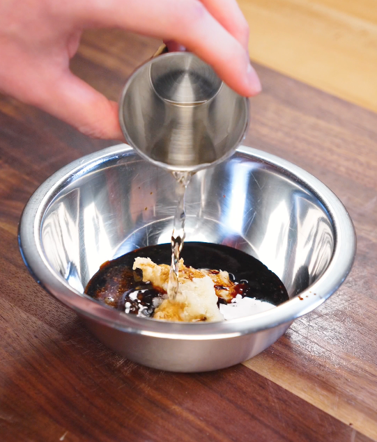 Rice vinegar being poured into a bowl with sesame paste, soy sauce, garlic, ginger, and sugar to make garlic sesame noodle sauce.