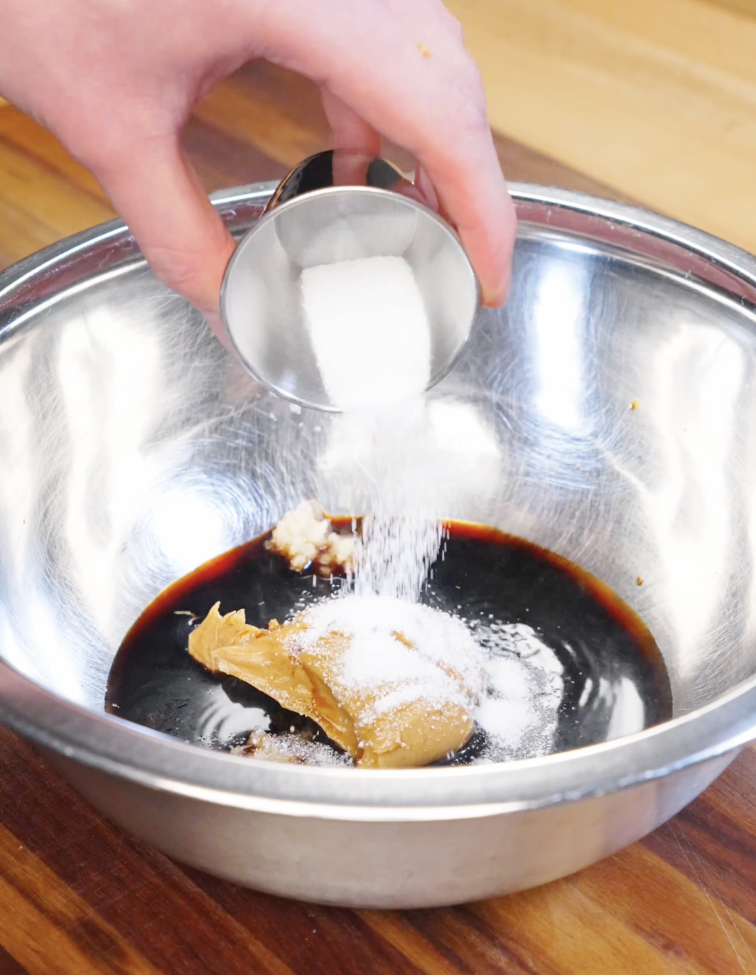 Sugar being added to a bowl with peanut butter, soy sauce, and garlic to make creamy peanut noodle sauce.