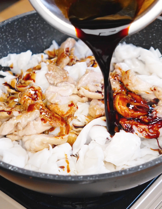 Dark glossy chow fun sauce being poured over wide rice noodles and chicken in a pan to coat everything evenly.