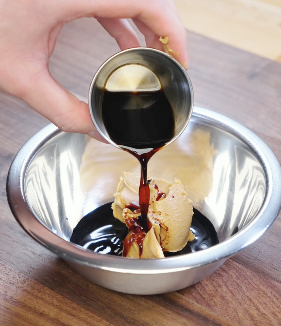 Dark soy sauce being poured into a bowl with creamy peanut butter to build the savory base for garlic peanut butter noodle sauce.