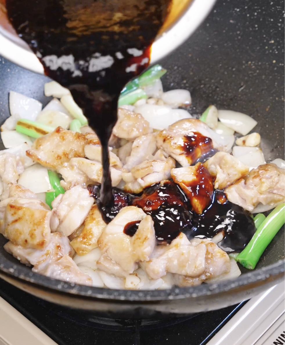 Thick honey garlic chicken stir fry sauce being poured over browned chicken, onions, and scallions in a hot pan.
