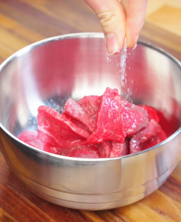 Hand sprinkling salt over thinly sliced beef in a metal bowl while preparing the marinade for Kung Pao Beef.