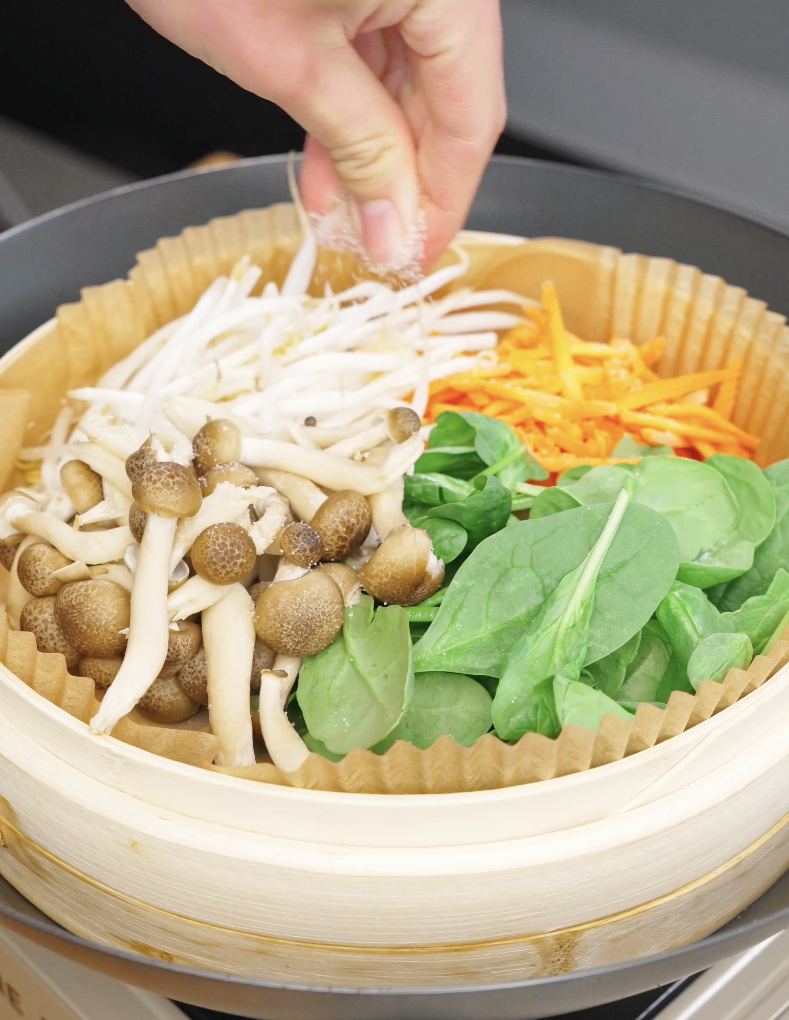 Hand sprinkling salt over mushrooms bean sprouts spinach and carrots in a steamer for beef bibimbap recipe.