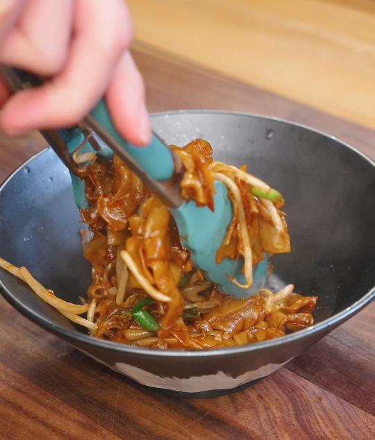 Silicone tongs lifting glossy chicken chow fun with wide rice noodles, bean sprouts, scallions, and tender chicken into a serving bowl.