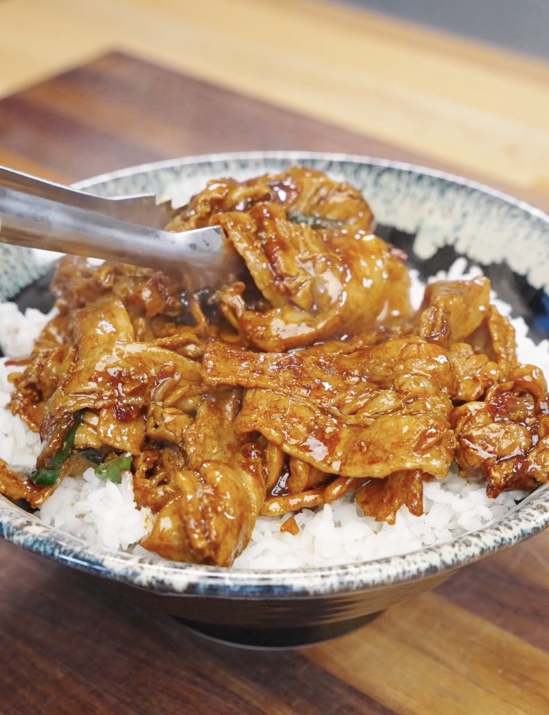 Garlic pork stir fry recipe being placed over a bowl of steamed white rice with sauce soaking into the grains.