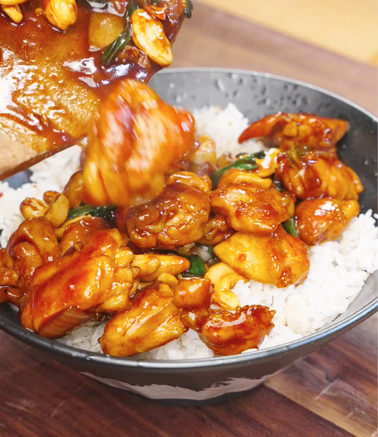 Honey cashew chicken with roasted cashews and scallions being spooned from a pan onto a bowl of steamed white rice.