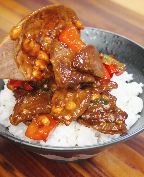 Wooden spoon placing glossy Kung Pao Beef with bell peppers and peanuts over a bowl of freshly steamed white rice.