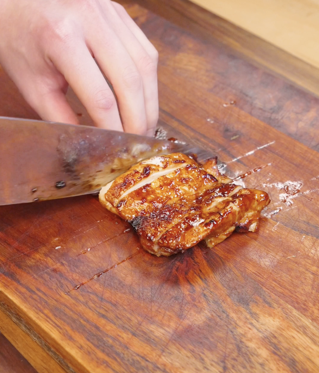 Knife slicing cooked Sweet Chili Chicken thighs into strips on a wooden cutting board.