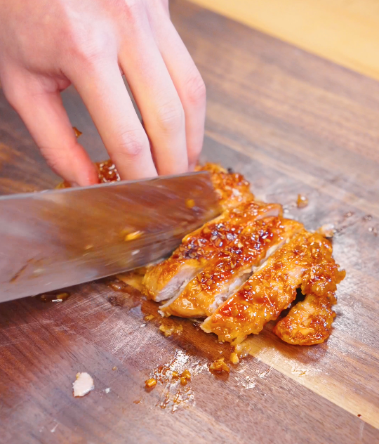 Knife slicing crispy garlic chicken into strips on a wooden cutting board after coating in garlic glaze.