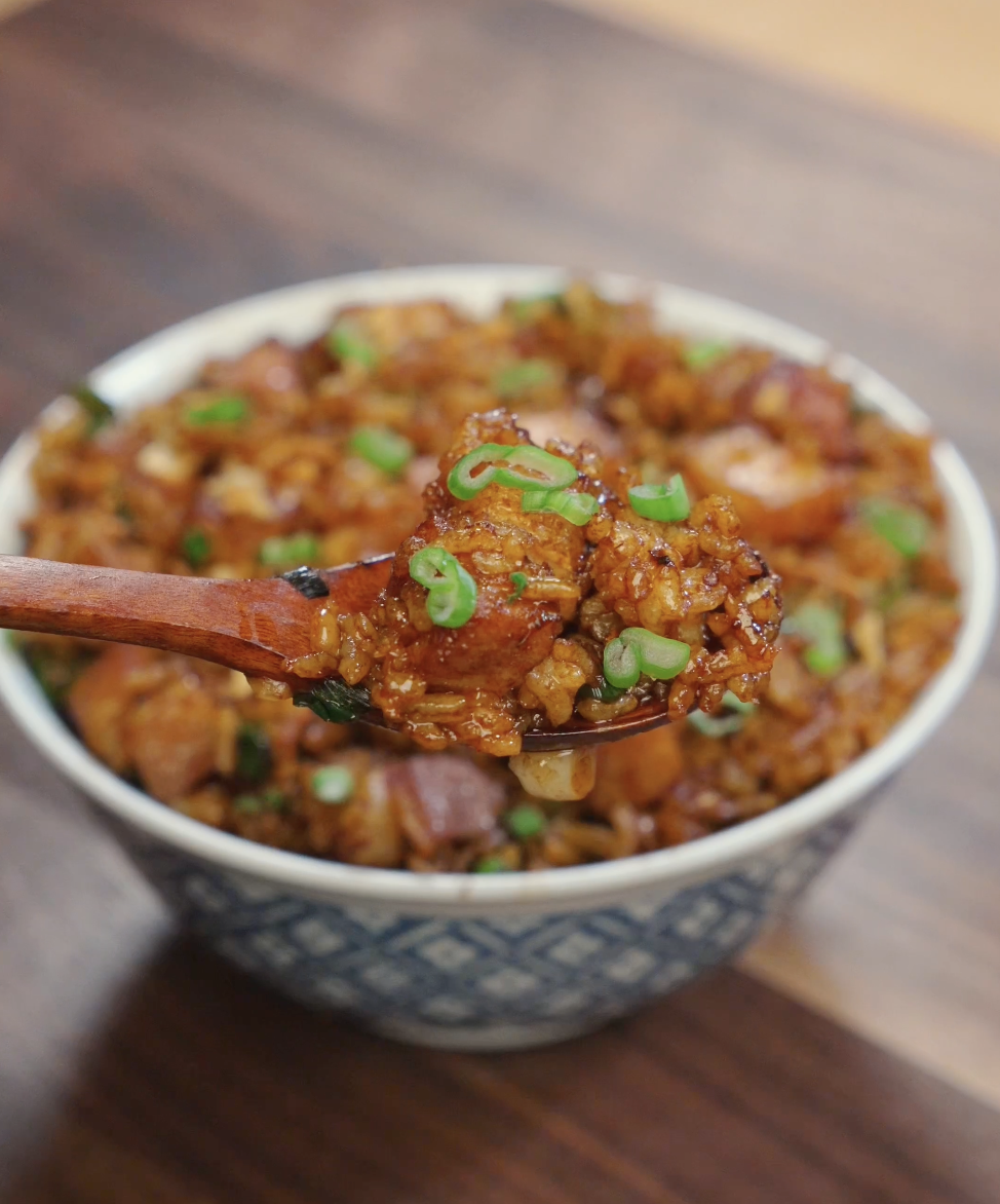 Wooden spoon lifting a bite of garlic pork belly fried rice with crispy pork pieces and scallions.