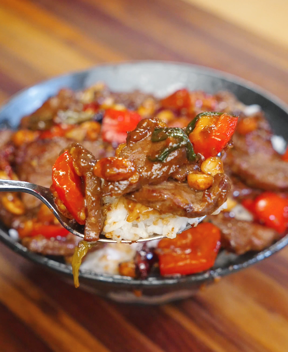 Fork lifting a bite of Kung Pao Beef with red bell peppers, peanuts, and glossy kung pao sauce over steamed rice.