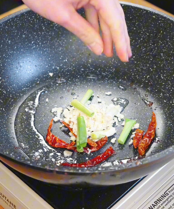 Minced garlic, dried red chilies, and scallion whites cooking in oil in a pan to build the aromatic base for Kung Pao Beef.