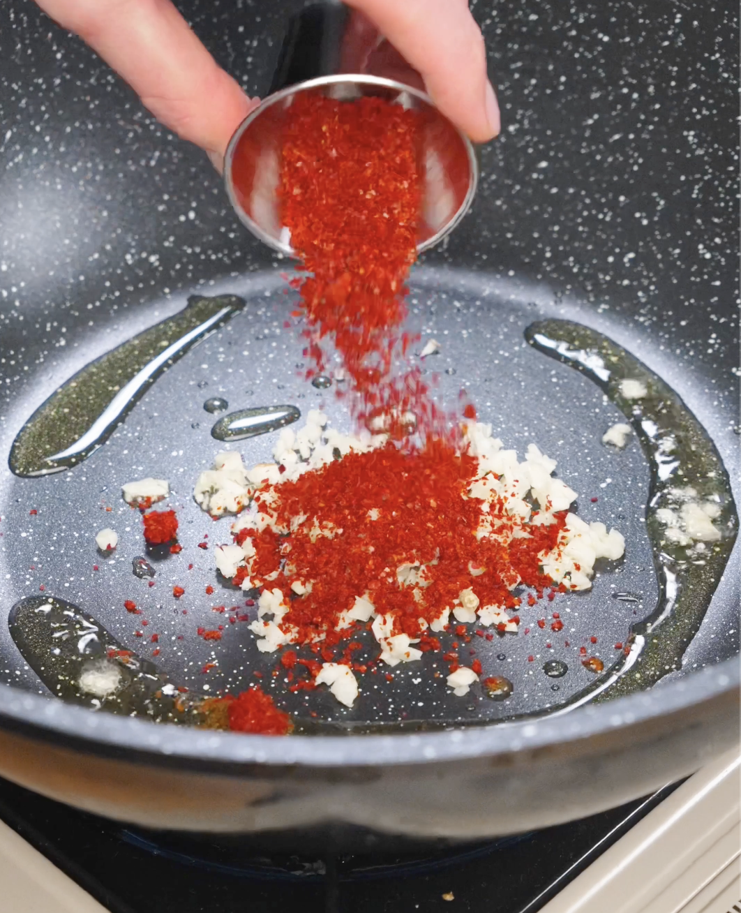 Chili flakes being poured into a pan with minced garlic and oil to begin making garlic chili noodle sauce.