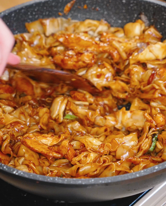 Wooden spatula tossing wide rice noodles, chicken, bean sprouts, and onions together in glossy chow fun sauce while stir frying.