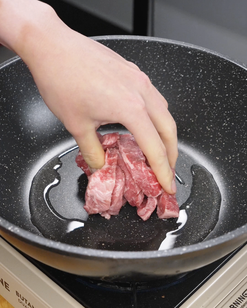 Hand placing marinated sliced beef into a hot pan with oil, starting the searing process for the Mongolian Beef Recipe.