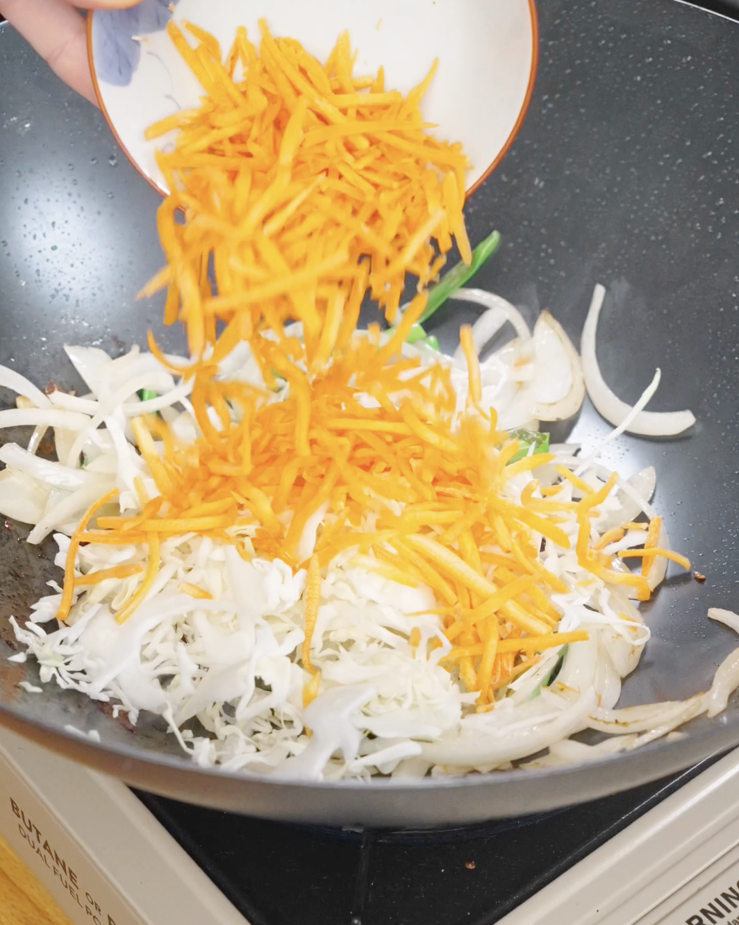 Shredded cabbage and carrots being added to saut&eacute;ed onions and scallions, adding texture and volume to chicken chow mein.