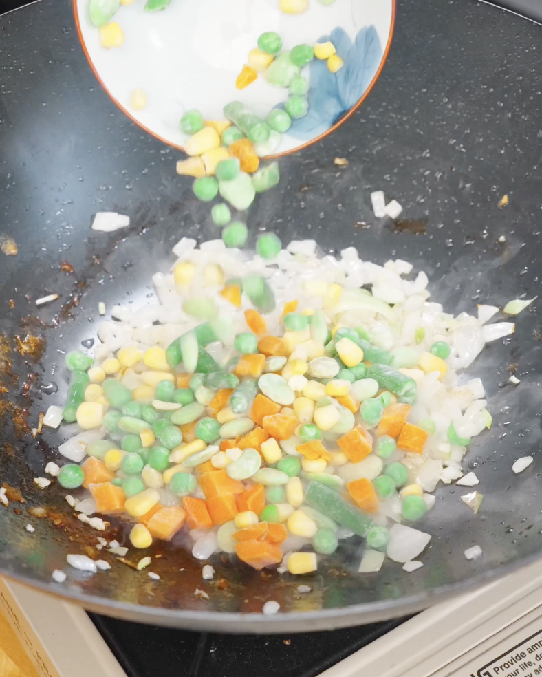Frozen mixed vegetables being added to saut&eacute;ed onions in a pan, adding color and texture to chicken fried rice.