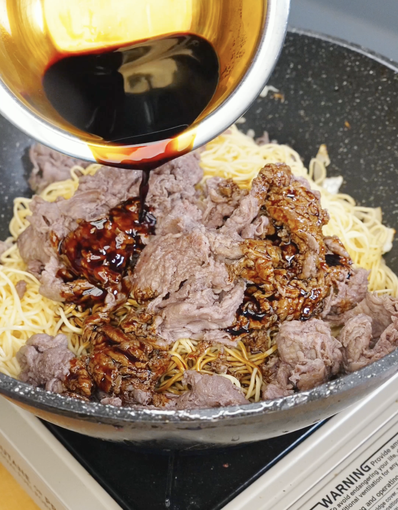 Cooked beef and chow mein noodles being tossed with dark soy sauce mixture in a pan for the Beef Chow Mein Recipe.