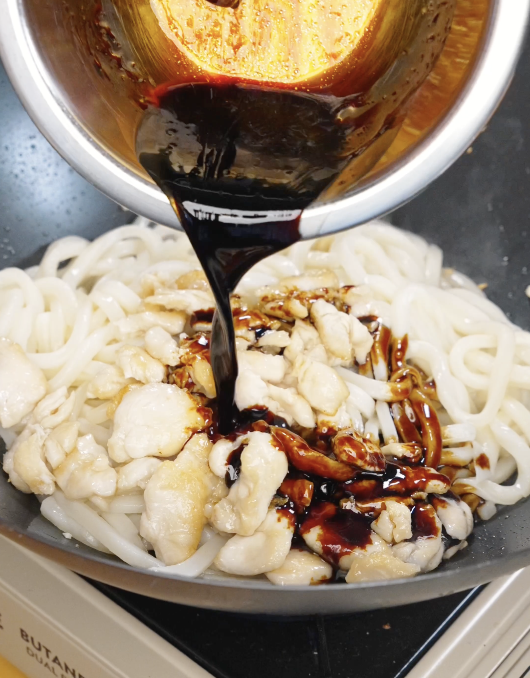 Sauce being poured over chicken and noodles in a pan for garlic chicken noodles.