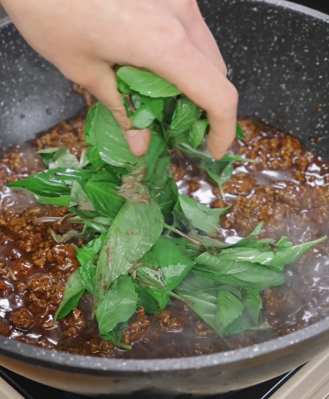 Fresh Thai basil leaves being added to saucy ground beef in a pan, infusing the dish with aromatic flavor.