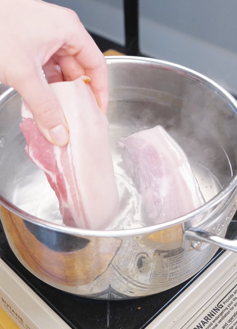 Raw pork belly being placed into a pot of boiling salted water to tenderize before crisping in the Vietnamese Crispy Pork Belly Recipe.
