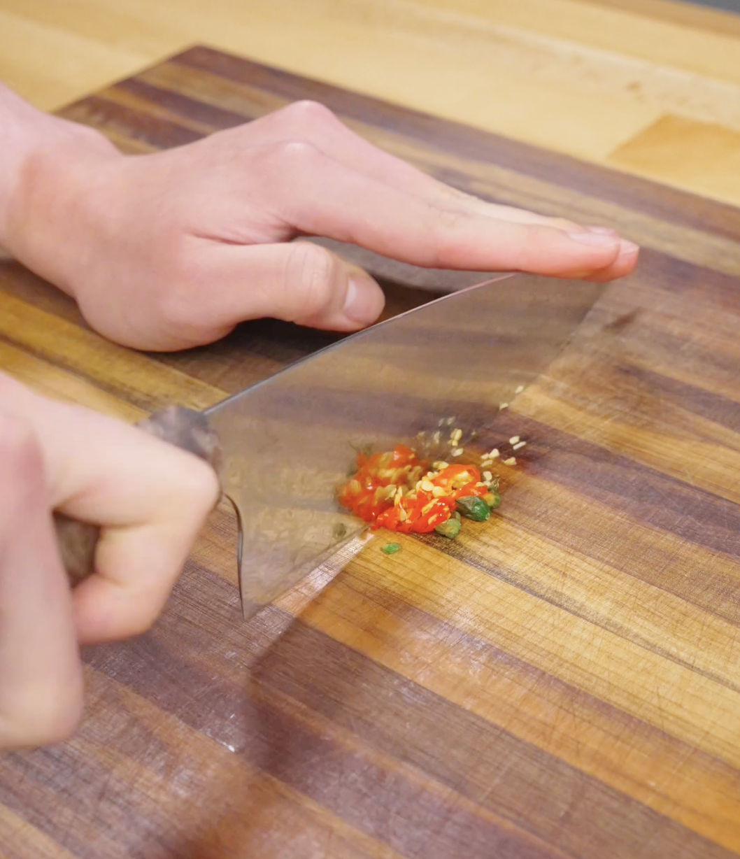 Fresh Thai chilies being finely chopped on a wooden cutting board, preparing the spicy aromatic base for Thai basil ground beef.