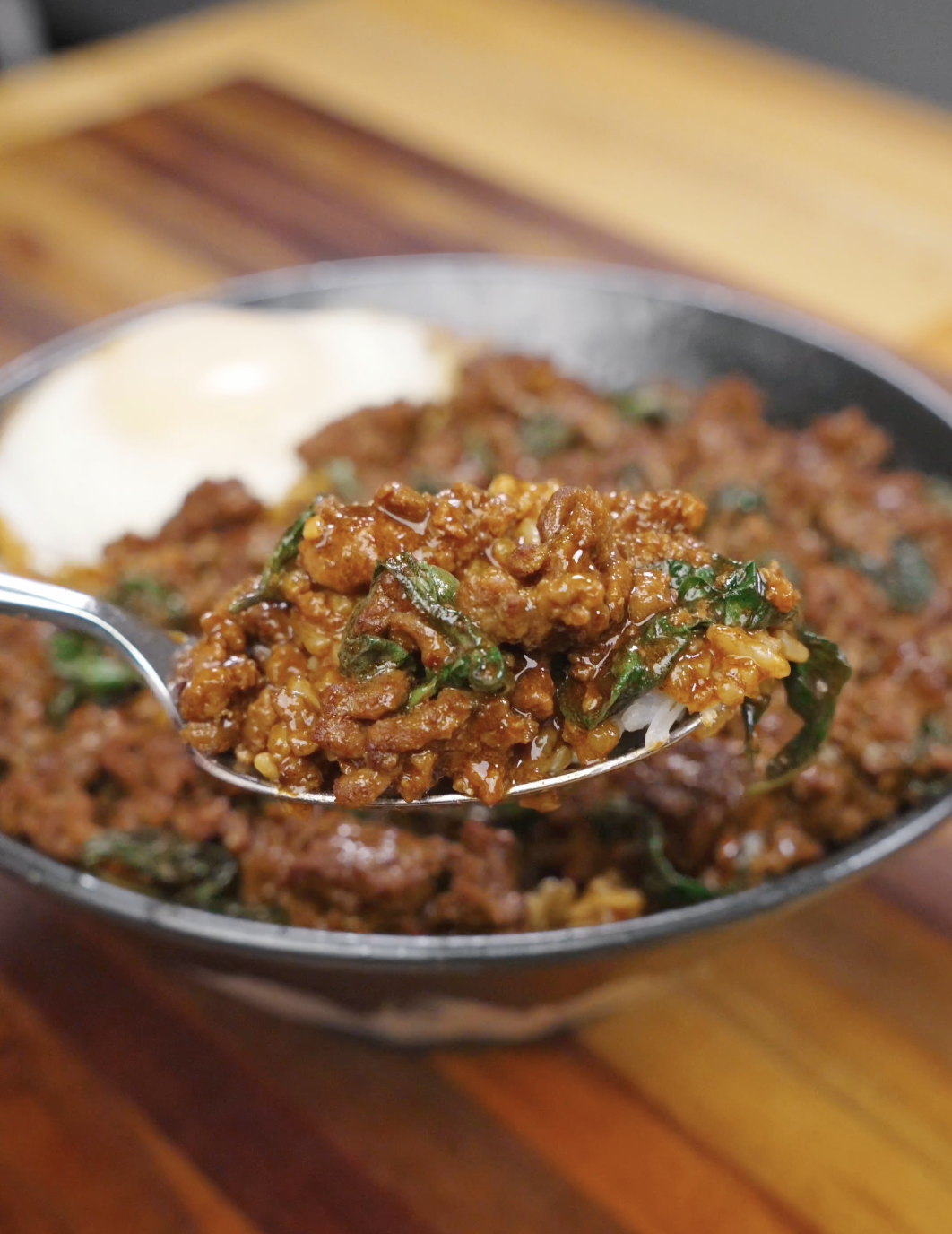 Spoon lifting glossy Thai basil ground beef over rice, showing caramelized sauce coating the meat and basil leaves.