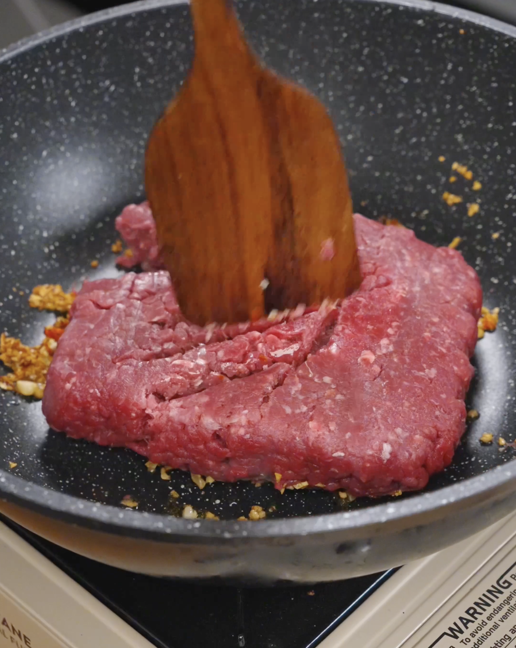 Ground beef being broken apart in a pan over aromatics, starting the browning process for Thai basil ground beef.
