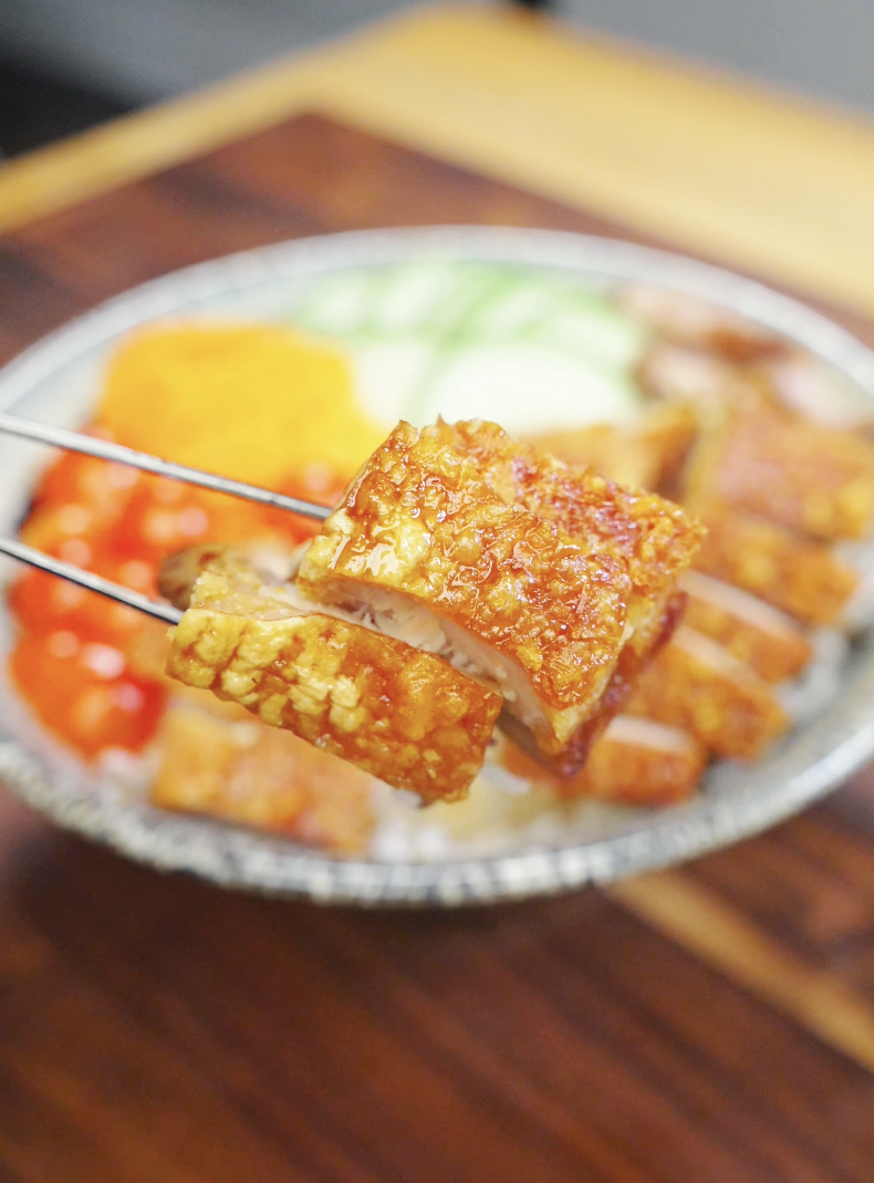 Chopsticks holding a piece of crispy pork belly showing golden crackling skin and juicy interior over a rice bowl in the Vietnamese Crispy Pork Belly Recipe.
