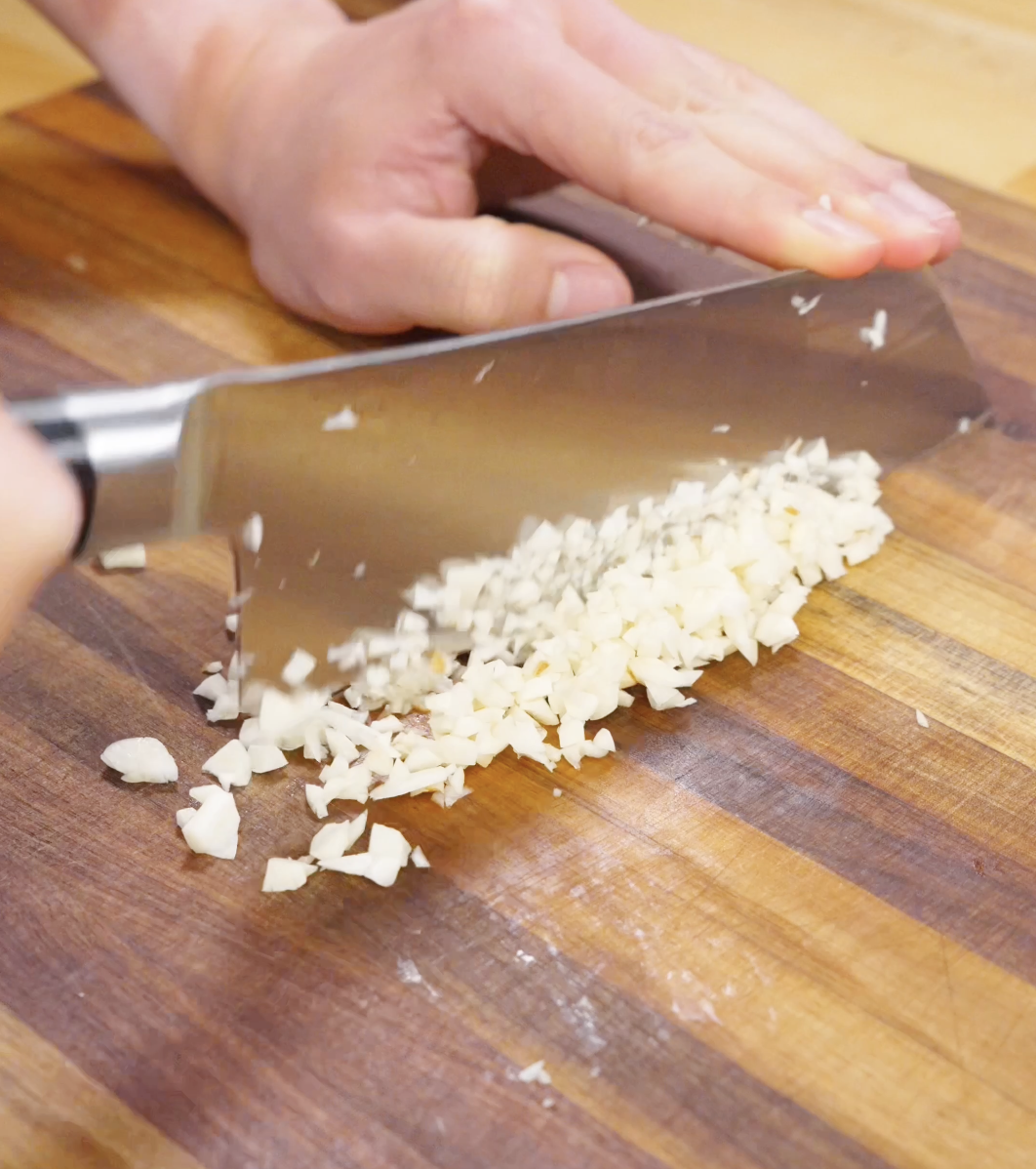 Garlic being finely minced on a wooden cutting board with a chef&rsquo;s knife, preparing aromatic base for garlic beef fried rice.