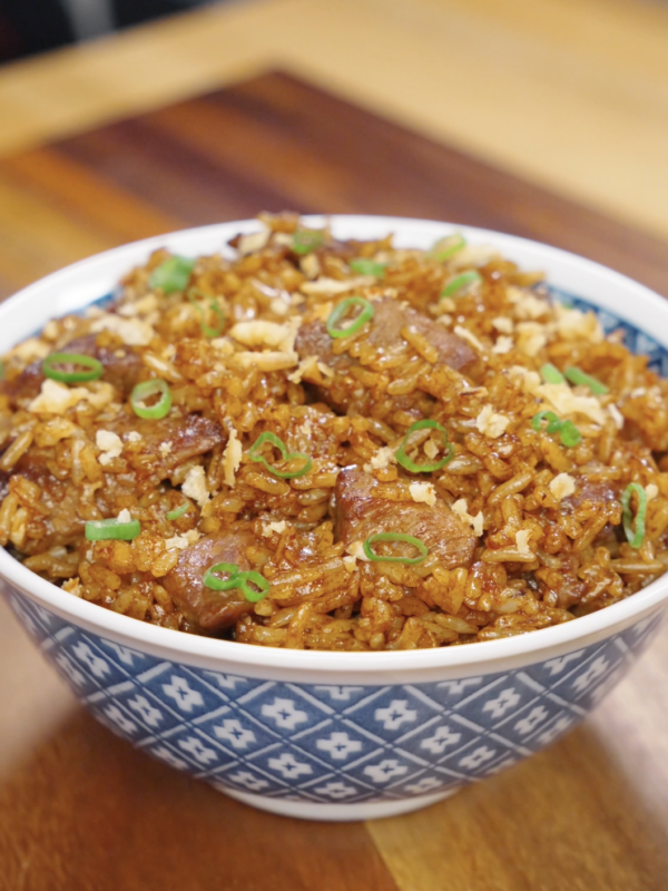 A bowl of garlic beef fried rice topped with crispy onions and sliced scallions, featuring caramelized beef and evenly coated rice with a deep savory glaze.