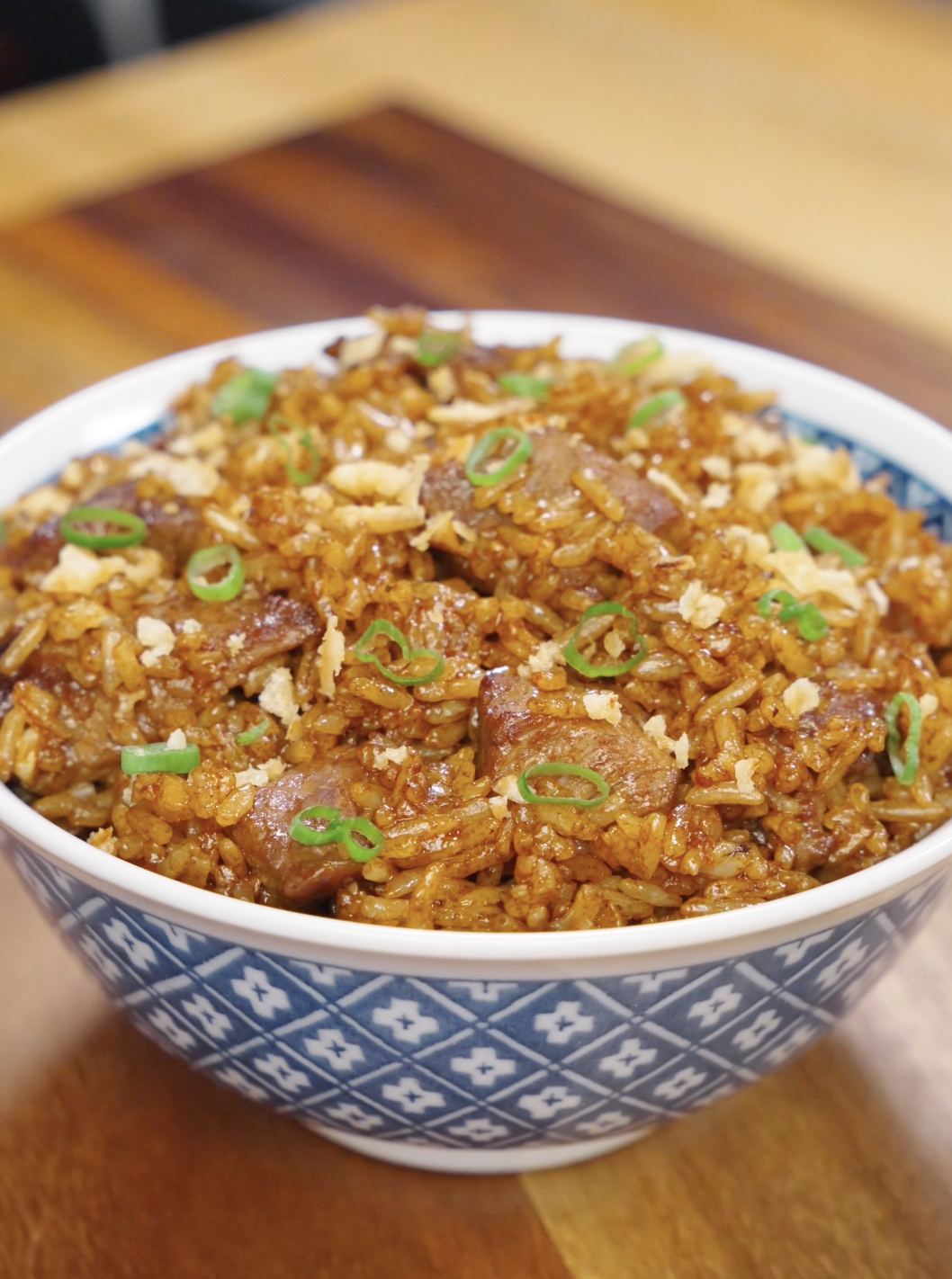 A bowl of garlic beef fried rice topped with crispy onions and sliced scallions, featuring caramelized beef and evenly coated rice with a deep savory glaze.