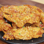 Crispy honey butter chicken tenders arranged on a plate, coated in a glossy sweet glaze and topped with sesame seeds and scallions.