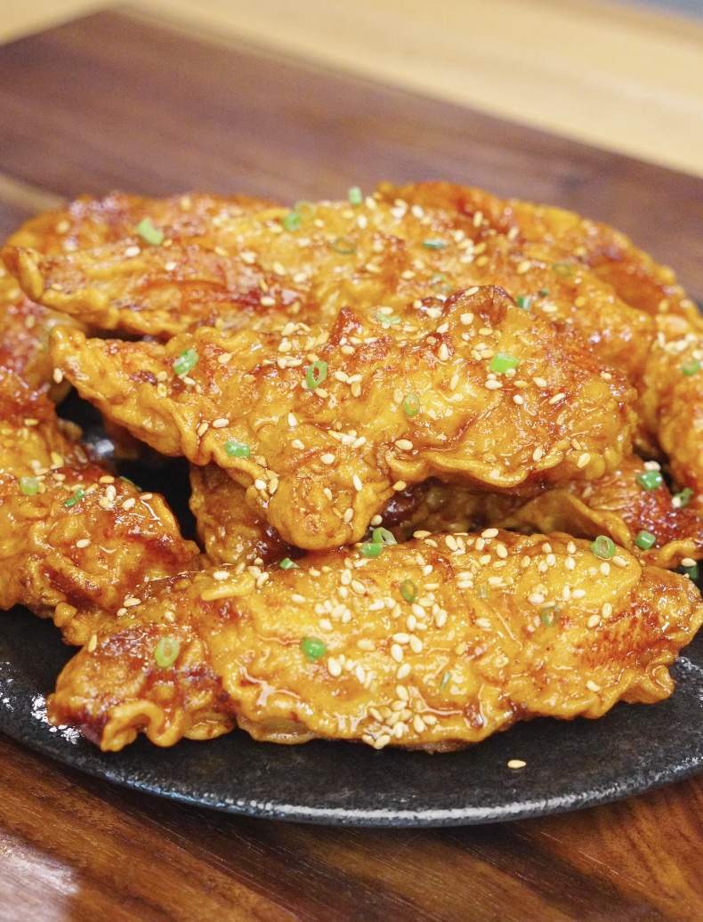 Crispy honey butter chicken tenders arranged on a plate, coated in a glossy sweet glaze and topped with sesame seeds and scallions.